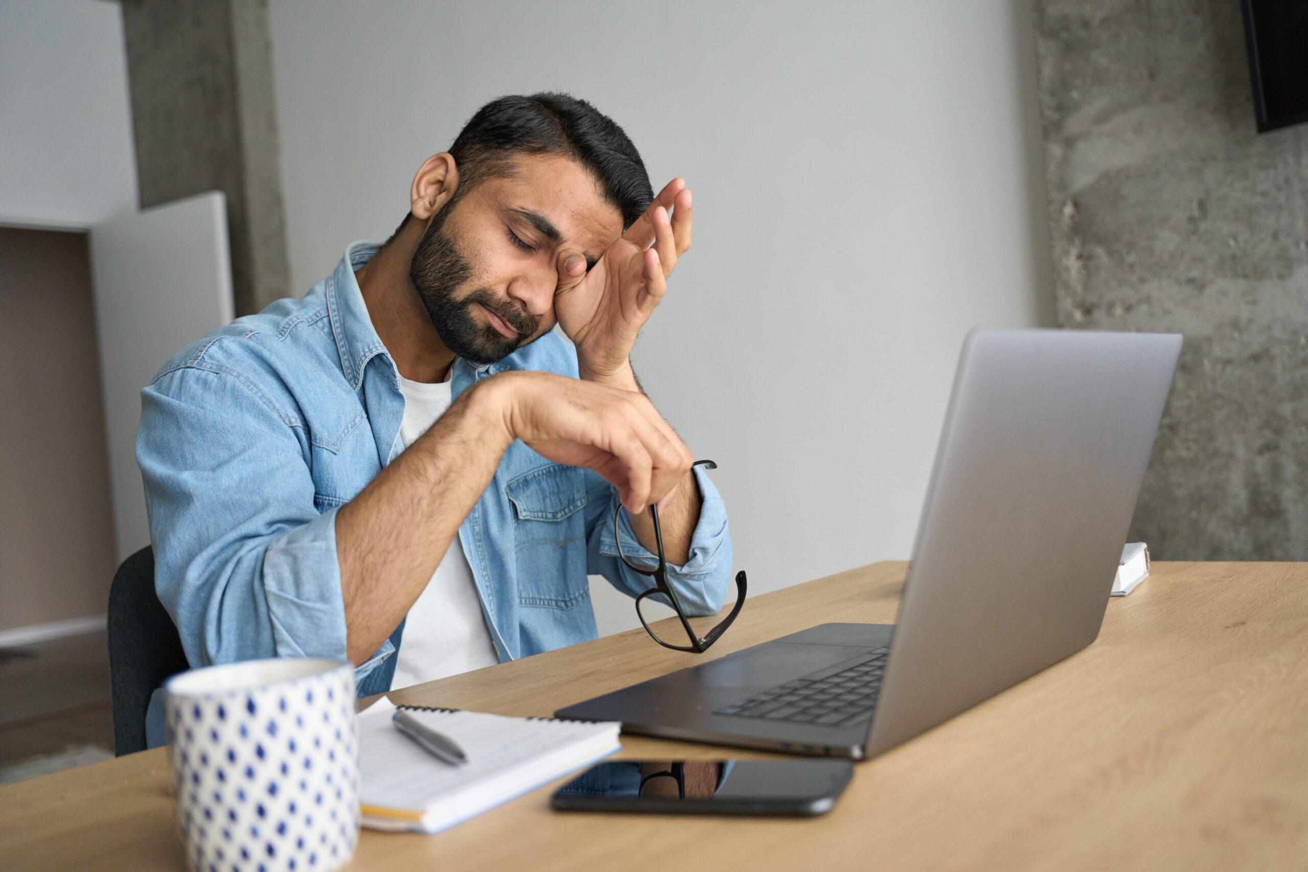Young indian eastern tired exhausted anxious business man rubbing eyes sitting in modern home office with laptop on desk. Overworked burnout student with glasses in hand feeling eyestrain.